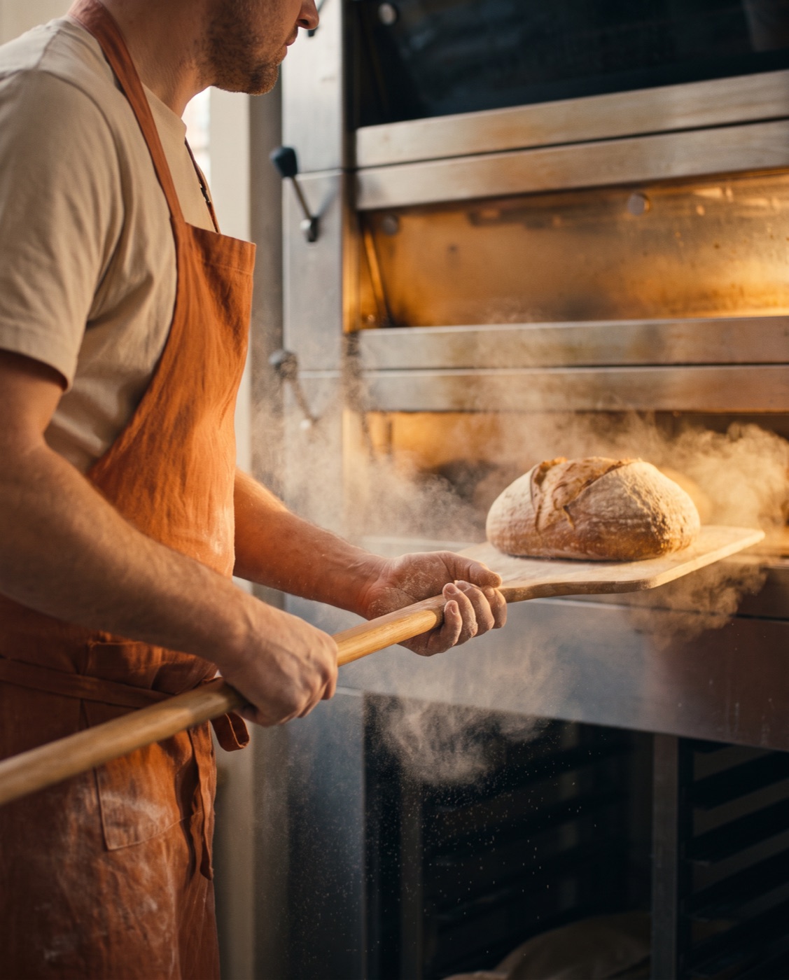 the baker slides a scored loaf on a wooden peel into the glowing deck oven