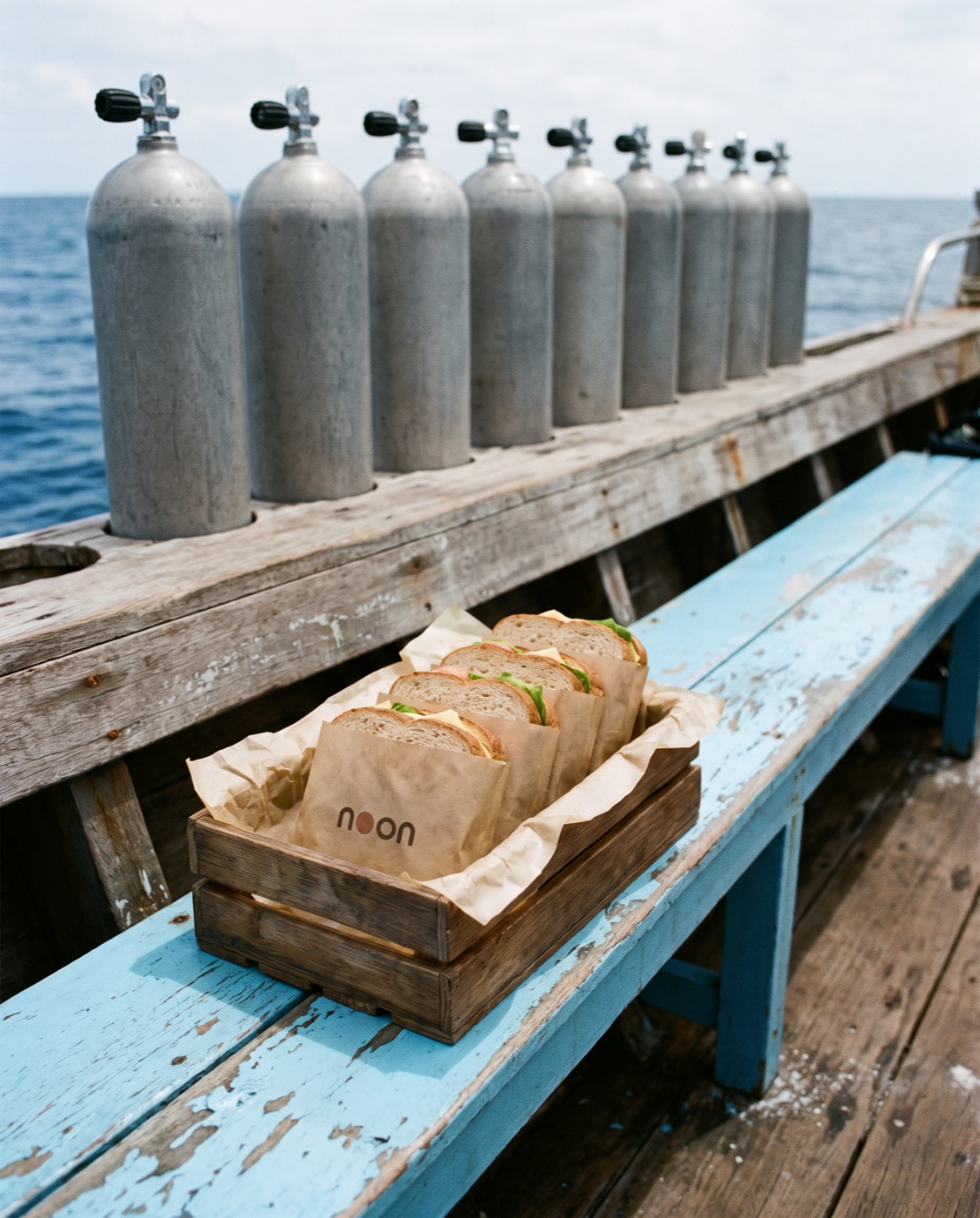 a crate of kraft-wrapped noon sandwiches on a dive boat bench, scuba tanks along the rail behind