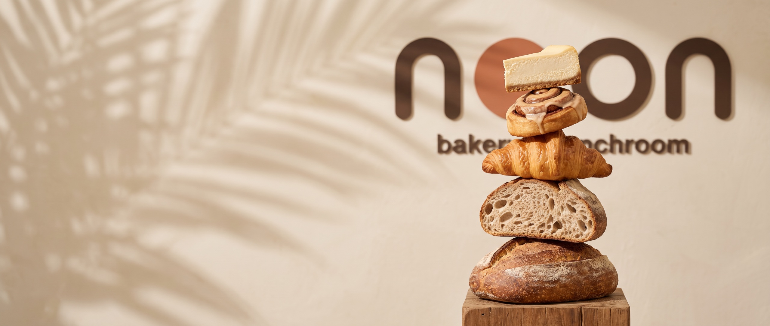 a tower of today's bakes — cheesecake, cinnamon roll, croissant and sourdough — in front of the noon wordmark, palm shadows on the limewash wall