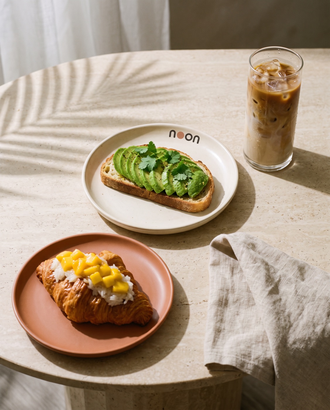 the regulars&rsquo; order from above: avocado toast on a noon plate, mango sticky rice croissant and an iced latte on travertine