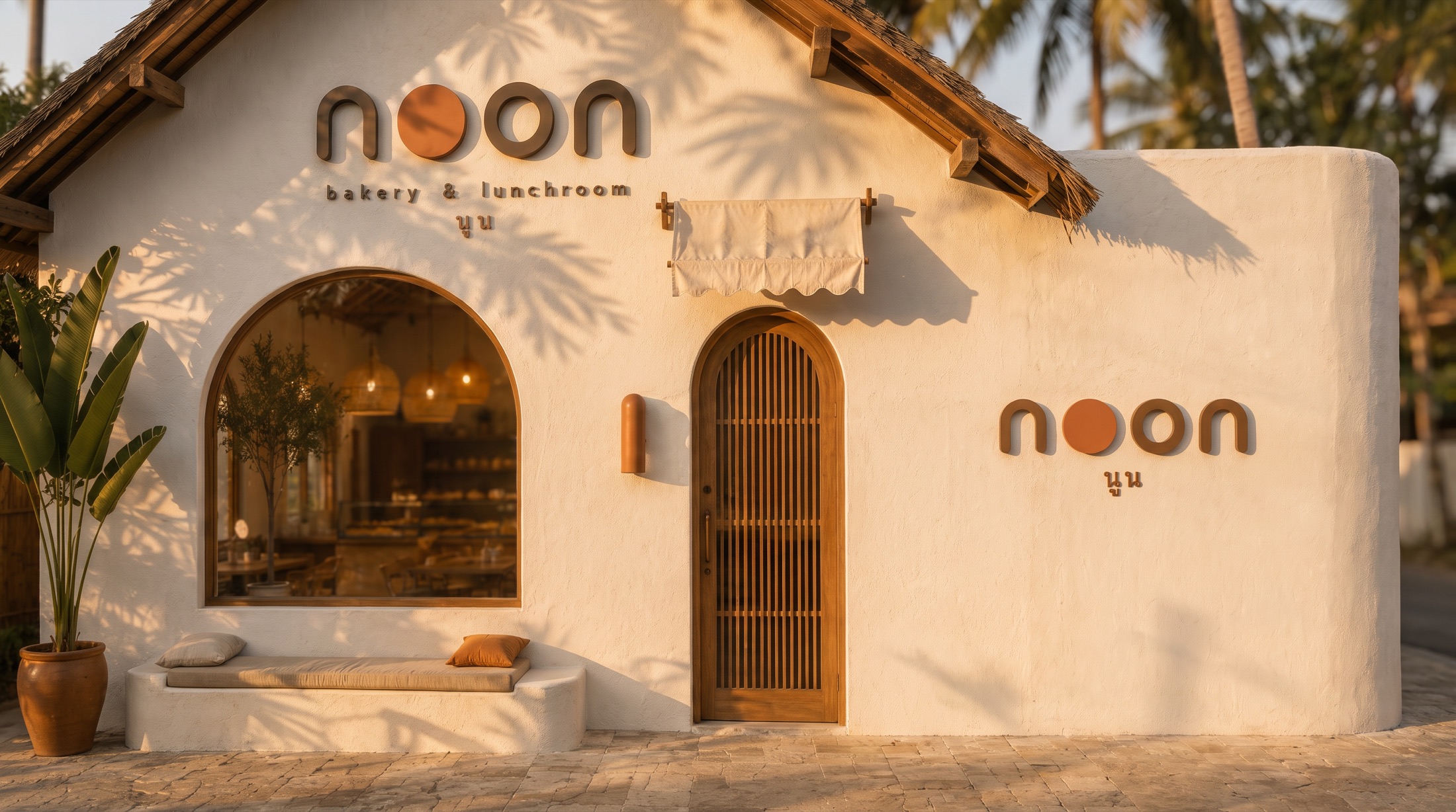 the noon facade in morning light: limewash gable, arched window with a built-in bench, arched teak door, palm shadows, the wordmark and นูน on the wall